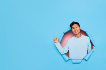 Smiling young man wearing light blue long sleeve standing on blue background pointing on the right...