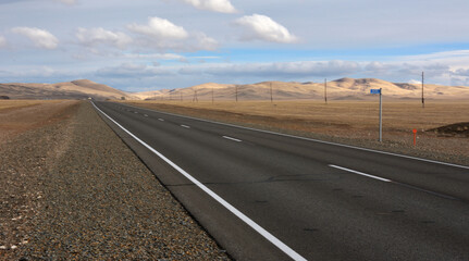 A new two-lane paved road with markings crosses the autumn steppe towards high hills under a cloudy overcast sky.