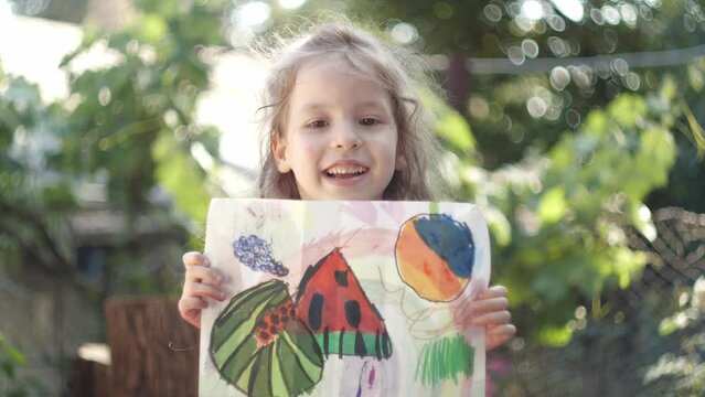 Portrait of a smiling child with a child's drawing. Creative development of the child, children's art school. Happy little girl holding a children's drawing outdoor. Summer sunny day