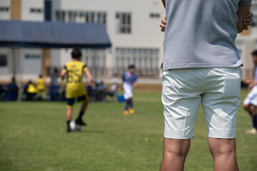 Father standing and watching his son playing football in a school tournament on a clear sky and sunny day. Sport, outdoor active, lifestyle, happy family and soccer mom and soccer dad concept.