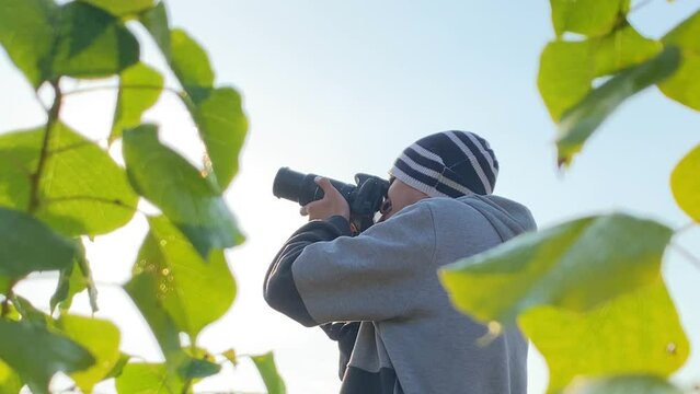 Man framed among leaves wears beanie and jacket as he takes pictures with telephoto lens