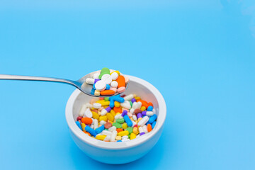 A mortar is placed on a blue background and filled with colorful pills capsules, a spoon and an empty space to use as a copyspace. Someone is floating with a spoon