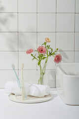 Beauty photo with bathroom concept. Toothbrushes in glass cup, white towel on round dishes, one corner of the washbasin and pink flower vase on tile wall background. Front view, copy space