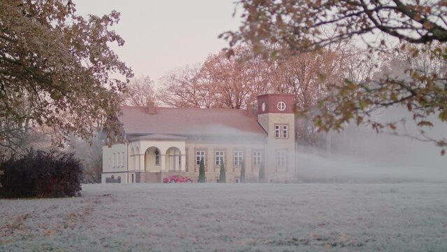 View to a luxury villa with a sleek sports car parked in front. Frosty and foggy early autumn morning during sunrise.