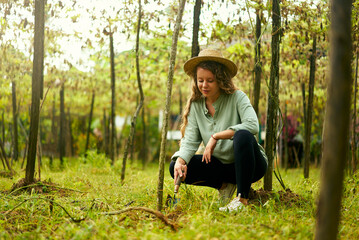 Young happy farmer woman in straw hat sitting in garden digging soil with garden shovel taking care...