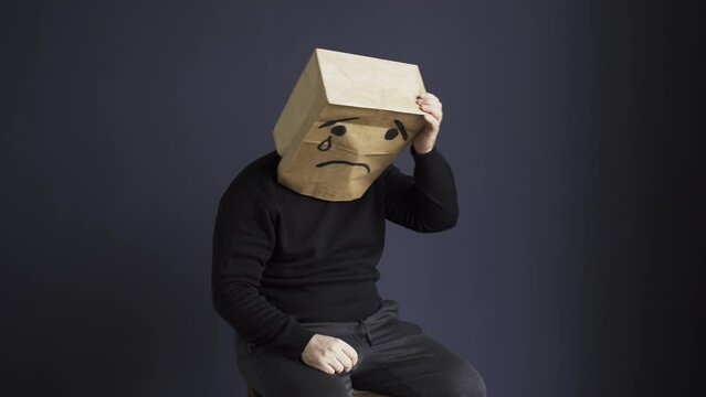 A sad man in a black turtleneck with a bag on his head, with a drawn crying emoticon, holds his head and cries. Emotions and gestures.