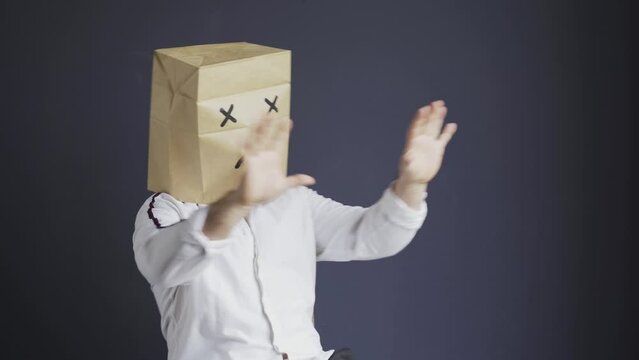 A sad man in a white shirt with a bag on his head, with a sad emoticon drawn, is afraid, defends himself by putting his hands forward. Emotions and gestures.