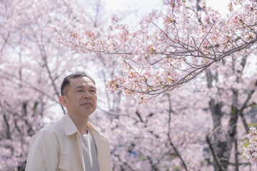 Middle-aged Asian man wearing white cardigan with cherry blossoms in background.