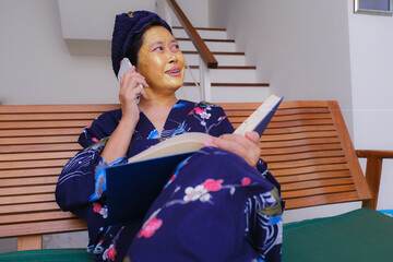 A woman with a beauty mask on her face is talking on the phone; smiling, an open book on her lap.