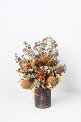 Beautiful dried flower arrangement of Australian native banksia, eucalyptus leaves, red leucadendrons and delicate white flowers, in a glass vase on a table with a white background.