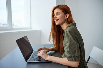portrait of a cute, smiling red-haired woman working at a laptop while sitting at an empty table in a bright room