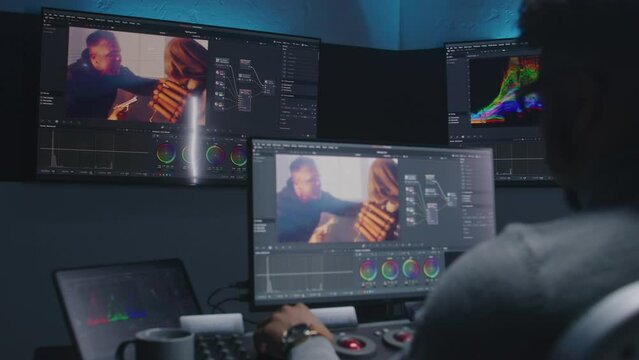 African American movie maker does color grading on computer in studio with equipment. Big monitors with action film footage and professional software interface on wall. Video post production process.
