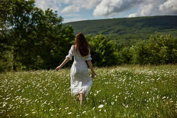 Fototapeta na wymiar a woman in a long light dress stands in a chamomile field against the backdrop of hills with her back to the camera enjoying nature and sunny weather