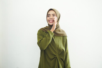 Beautiful young Asian woman in green dress and hijab hand on mouth telling secret, whispering gossip, looking at camera with cheerful expression on white background. People islam religious concept