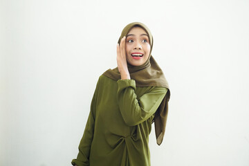 Beautiful young Asian woman in green dress and hijab hand on mouth telling secret, whispering gossip, looking at camera with cheerful expression on white background. People islam religious concept