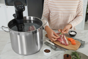 Woman adding rosemary to meat near pot with sous vide cooker at table, closeup. Thermal immersion...