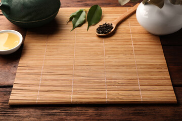 Bamboo mat, teapot, cup and spoon of dry tea leaves on wooden table, space for text