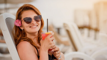 Young beautiful woman wearing sunglasses is lying on a beach deck chair and drinking a cocktail near a tropical sea.