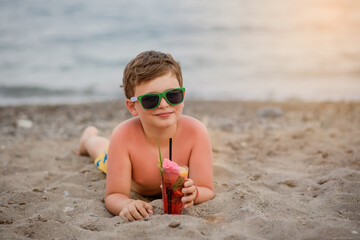Little boy kid with sunglasses is lying on the sand near seaside smiling and drinking a cocktail.