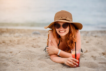 Young beautiful woman wearing sunglasses and a hat is lying on beach sand and drinking a cocktail near a tropical sea.