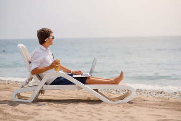 Young man in sunglasses and white shirt is lying on a beach chair on the seaside holding a cocktail and a laptop.