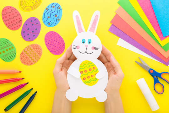Young adult woman hands with white smiling bunny and colorful eggs from paper on bright yellow table background. Point of view shot. Closeup. Making easter decoration elements. Top down view.
