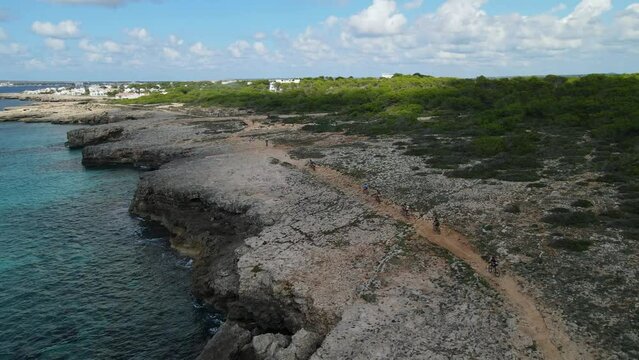 Cyclists ride along the coast line in Menorca with crystal clear blue waters seen as drone pans around riders.