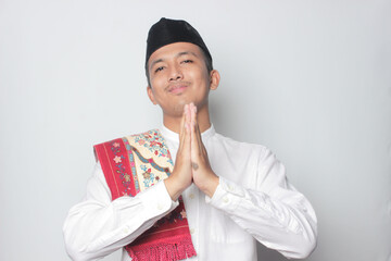 Smiling young Asian man wearing muslim shirt gesturing traditional greeting isolated on white background.