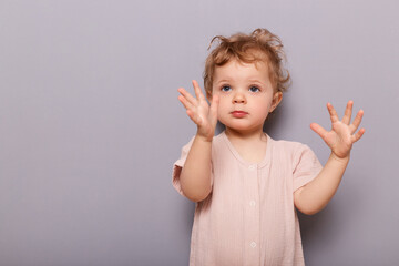 Portrait of calm focused toddler girl standing isolated over gray background looking away, raised her hands, looking at something interesting, copy space for advertisement.