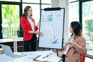 Two young business women meet to analyze the financial chart at the office to discuss the financial situation at the company. A partner sits at a desk with modern documents and equipment.