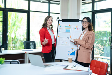 Two young business women meet to analyze the financial chart at the office to discuss the financial situation at the company. A partner sits at a desk with modern documents and equipment.
