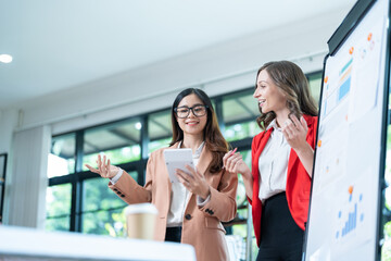 Two young business women meet to analyze the financial chart at the office to discuss the financial situation at the company. A partner sits at a desk with modern documents and equipment.