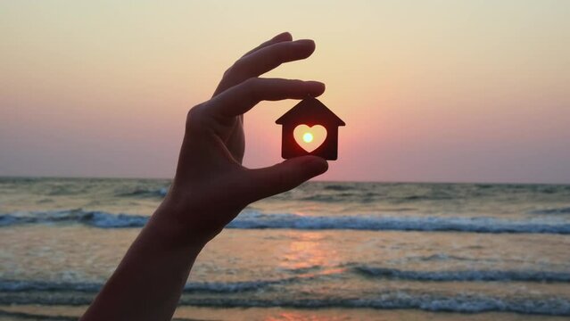 Close-up shot of human hand holding wooden house model on sunset and sea background. Seaside dream house concept. Choosing place for a new home.