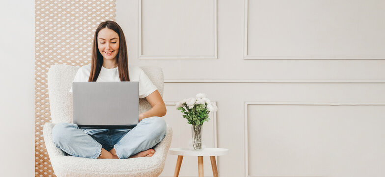 Young Woman Sits In A Chair And Works With A Laptop At Home Against The Background Of A White Wall