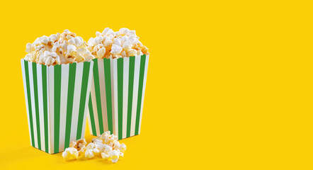 Two green white striped carton buckets with tasty cheese popcorn, isolated on yellow background. Box with scattering of popcorn grains. Fast food, movies, cinema and entertainment concept. - obrazy, fototapety, plakaty