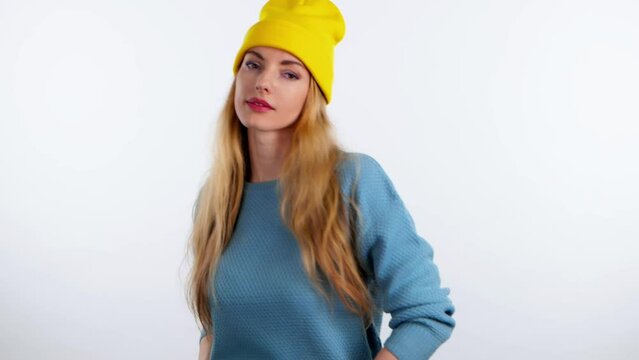 Cheerful young woman dancing. Funny smiling overjoyed blonde lady waving hands in yellow beanie and blue sweater. Indoor studio, emotional 20s girl enjoys dancing isolated on white background.