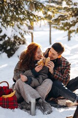 A young couple spends a day off at a picnic in the winter forest. Smiling young people sit in the middle of a winter forest and eat pancakes.