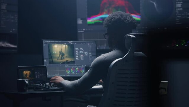 African American film editor does color grading on computer in modern studio. Big monitors with RGB colour correction graphic bar and program interface on the wall. Video post production. Handheld.
