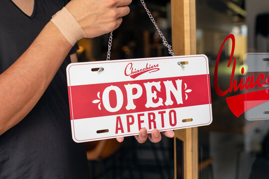 Man hanging up an Open sign on a restaurant door. Dual language, Italian and English.
