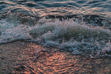 Wave coming on bank sea shore lit by evening sun. Water drops splashing. Close up of ocean sea lake wave.