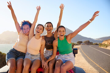 Portrait Of Laughing Female Friends Sitting On Hood Of Open Top Car On Road Trip