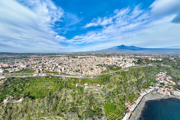 Fototapeta premium Acireale e la Timpa - Panoramica aerea dall'alto sulla Timpa di Acireale e sul porto di Santa Maria la Scala con vulcano Etna sullo sfondo del cielo blu con nuvole 