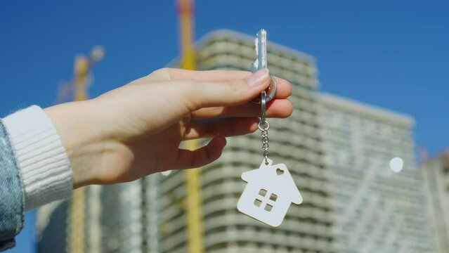 Hand with keychain in shape of small house and keys. Against backdrop of typical American country-style house. Construction, sale or purchase of apartments. Developer, realtor or real estate agency.