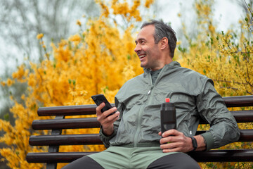 A middle shot of a fit middle-aged man taking a break from jogging and using his smartphone on a bench in the park.