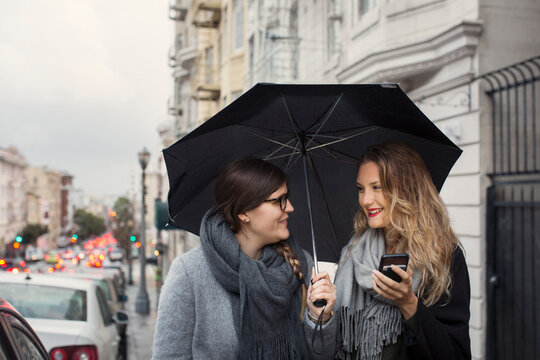 Two women sharing umbrella and using smart phone
