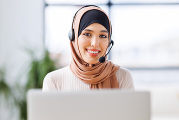 friendly smiling muslim woman with headset working in front of laptop talking while calling client