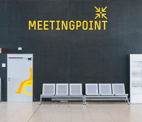 Yellow meeting point sign on a black wall next to some chairs to rest inside a station