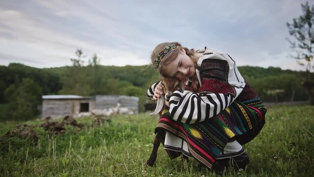 National costume of a Ukrainian young girl hugging a little goat on a green grass on a summer day