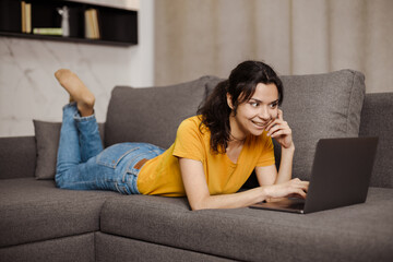Fototapeta premium Happy indian or arabian young woman lying sofa and using laptop. Remote working