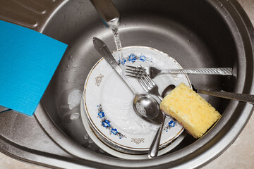 Washing dishes. Forks, spoons, plates of various sizes, rags and a sponge in the sink under running...